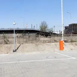 Podniesiona szlabanowa zapora parkingowa z pomarańczową obudową, widok na szary brukowany plac, ogrodzenie z drutu kolczastego i budynki przemysłowe w tle pod błękitnym niebem.