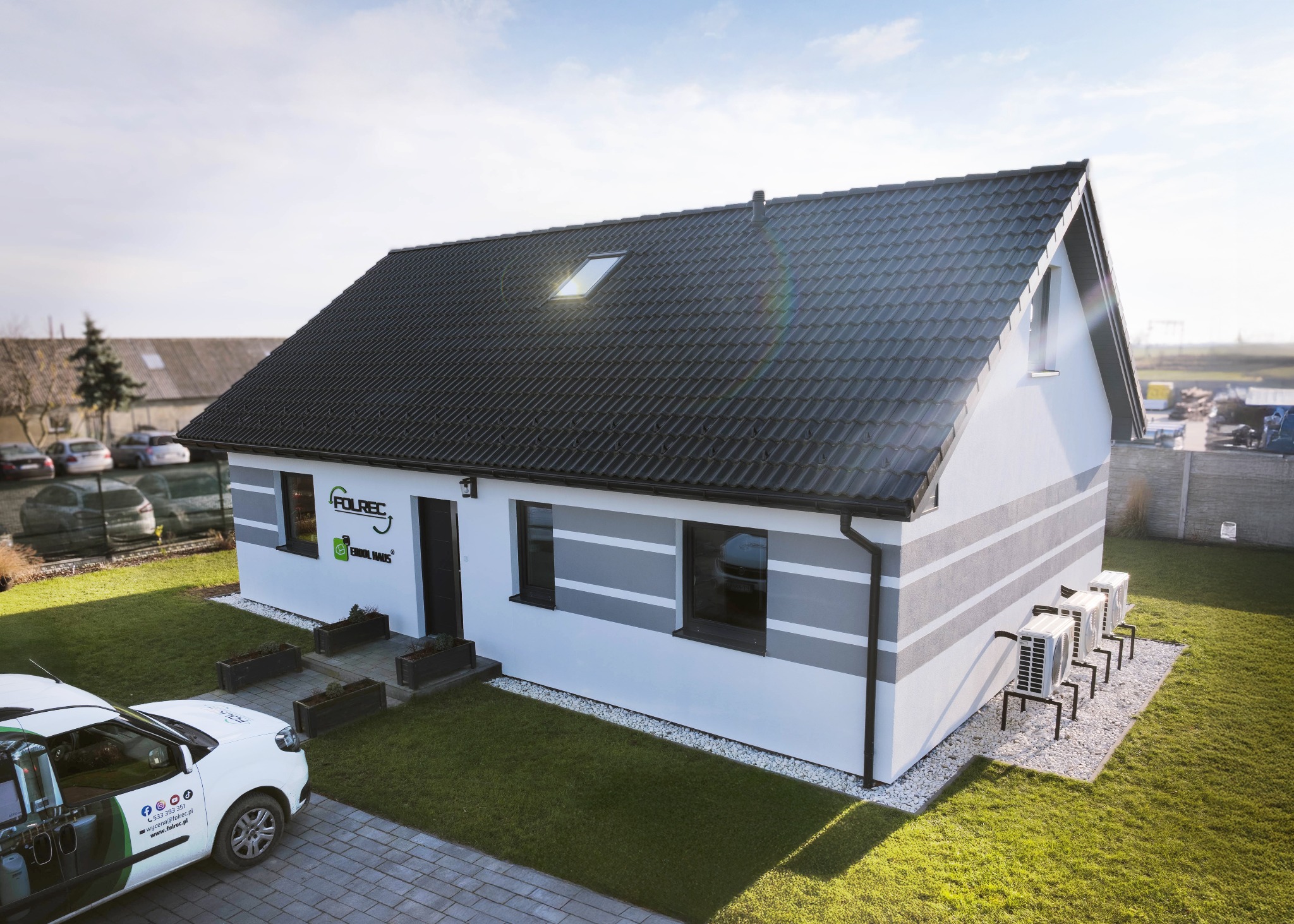 Gotowy dom szkieletowy z szarą elewacją i klimatyzacją, prezentowany z zewnątrz na tle zielonego trawnika i błękitnego nieba. Widoczny firmowy samochód.