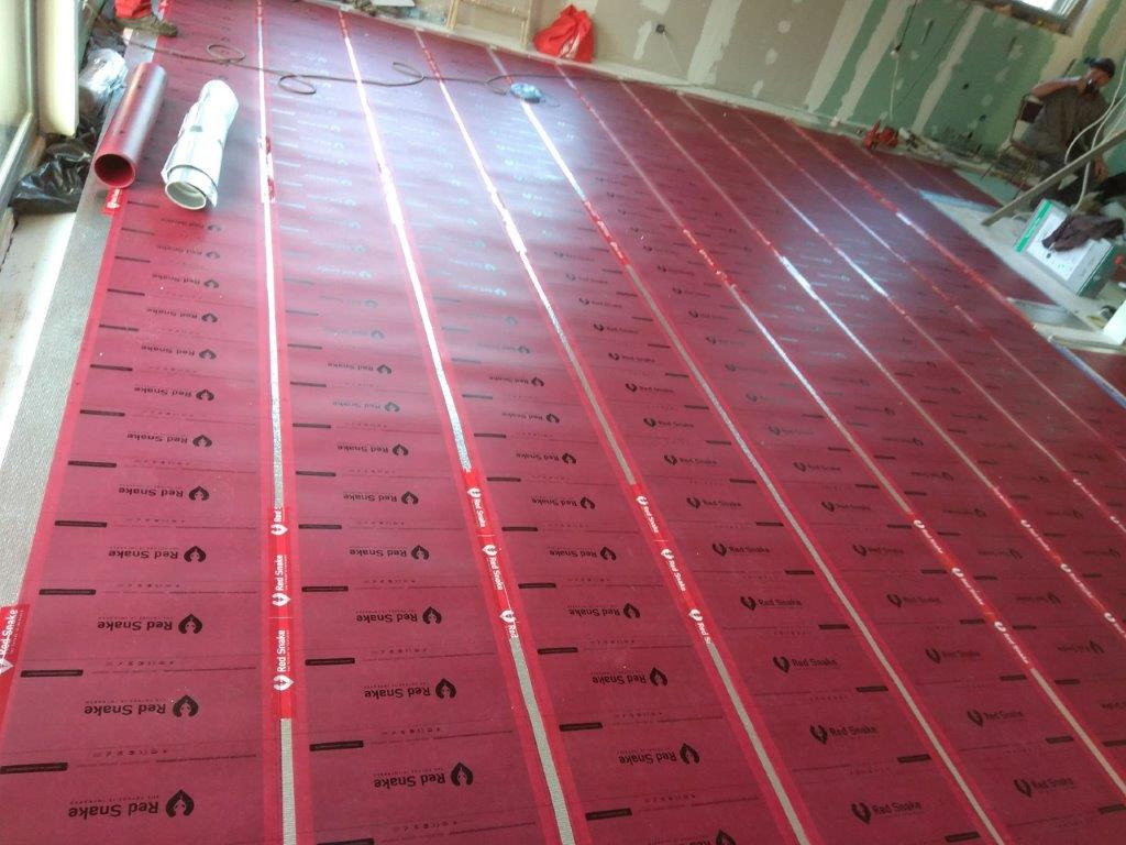 Rozłożone czerwone panele podłogowe z logo 'Red Snake' i srebrnymi paskami, przygotowane do montażu instalacji grzewczej w nowym budynku, widoczne rury i prace wykończeniowe w tle.