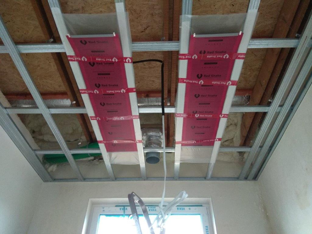 Widok instalacji pod sufitem podwieszanym z widocznymi kanałami wentylacyjnymi zabezpieczonymi folią Red Snake, rury wentylacyjne i fragment okna VEKA w tle.