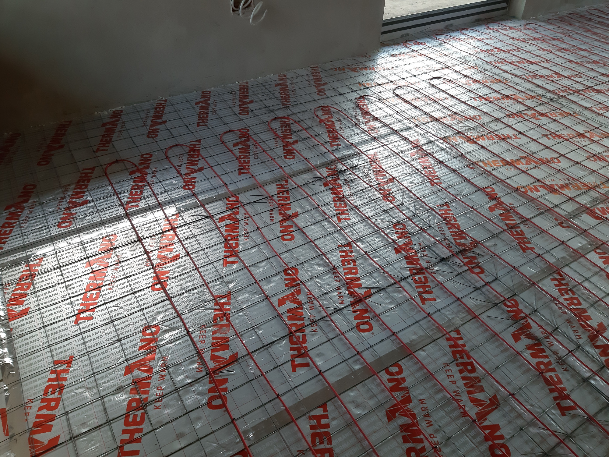 Rozłożone czerwone kable grzewcze Devi na srebrnej folii izolacyjnej THERMANO z nadrukowanym logo, przygotowane do zalania wylewką pod ogrzewanie podłogowe.