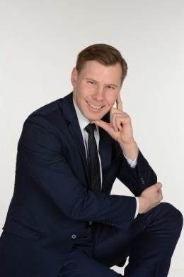 Uśmiechnięty mężczyzna w granatowym garniturze i czarnym krawacie, z dłonią przy policzku, na jasnym tle.