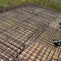 Zbrojenie płyty fundamentowej na deskowaniu, widok z góry. Na pierwszym planie cegła z zapiskami. W tle łąka i zabudowania w oddali.