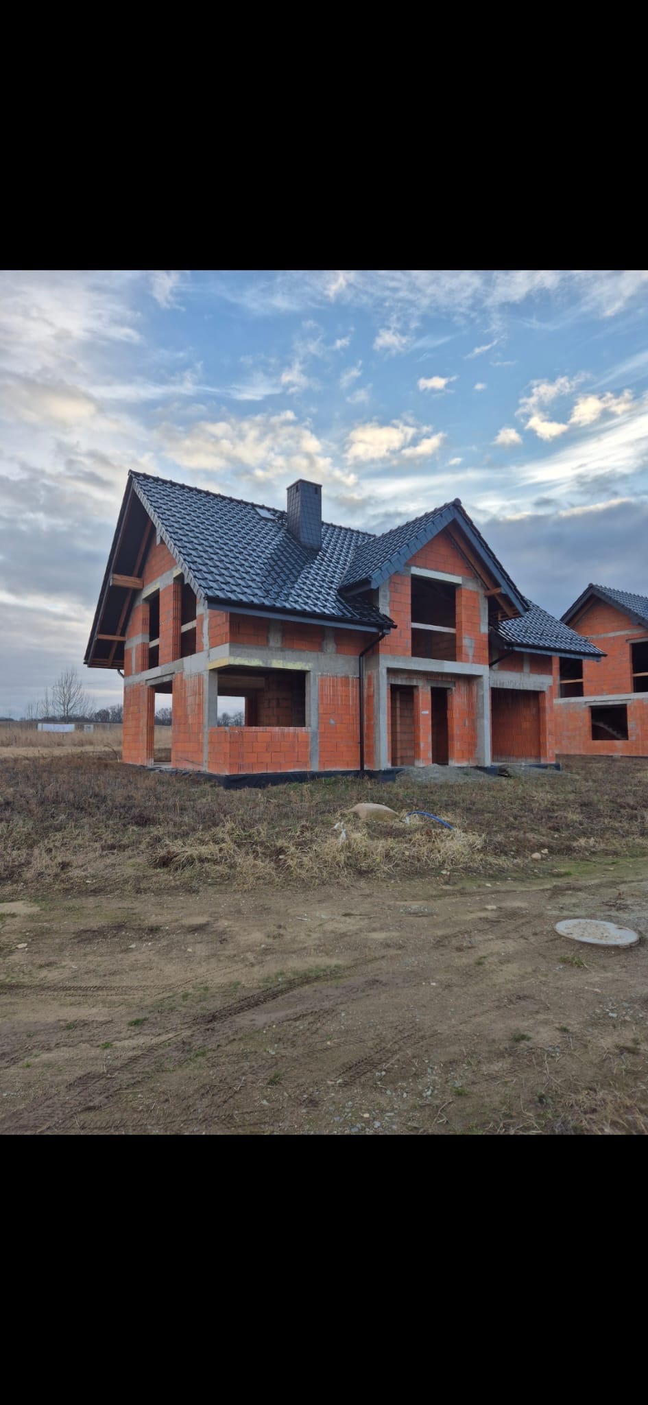 Budowa domu z czerwonej cegły, z ciemnym dachem, na tle pochmurnego nieba. Widoczne surowe ściany i otwory okienne. Stan surowy otwarty.