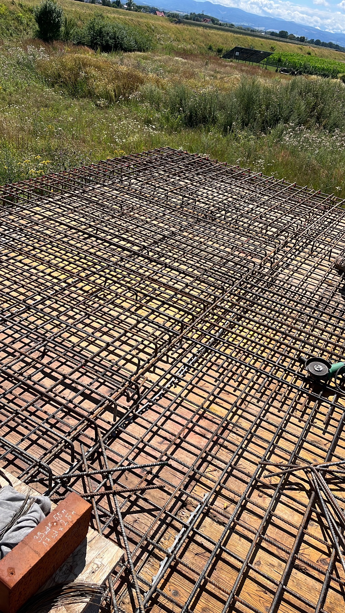 Zbrojenie płyty fundamentowej na deskowaniu, widok z góry. Na pierwszym planie cegła z zapiskami. W tle łąka i zabudowania w oddali.