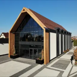 Nowoczesny budynek z drewnianą konstrukcją szkieletową, przeszkloną fasadą i brązową dachówką, otoczony kostką brukową w geometryczny wzór.