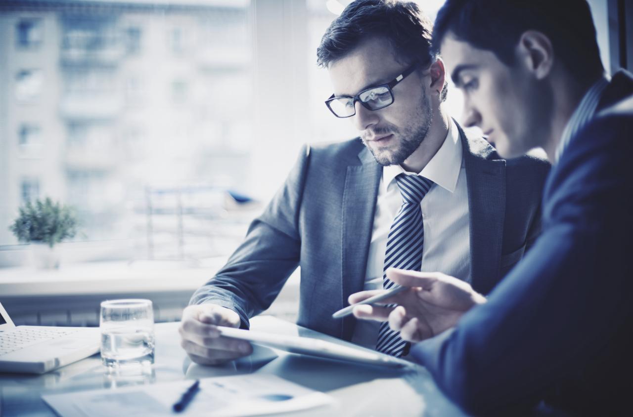 Dwaj mężczyźni w garniturach analizują dane na tablecie w jasnym biurze, widoczny laptop, szklanka wody, okno z rozproszonym światłem.