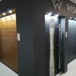 Wystawa bram garażowych firmy Wiśniowski: brama segmentowa w kolorze drewna z poziomymi panelami oraz brama w kolorze antracytowym z elementami ze stali nierdzewnej i przeszkleniem.