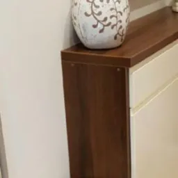 Fragment szafki na buty z ciemnobrązowym blatem i białymi frontami, na blacie stoi dekoracyjna, kremowa waza z brązowymi wzorami roślinnymi.