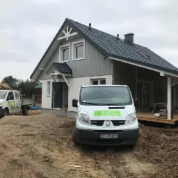 Dwukondygnacyjny dom z szarą elewacją i ciemnym dachem w trakcie budowy, z widocznymi dwoma samochodami dostawczymi firmy budowlanej na piaszczystym terenie.