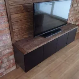 Czarna szafka RTV z drewnianym blatem pod telewizorem, na tle ściany z drewnianych paneli i cegły.