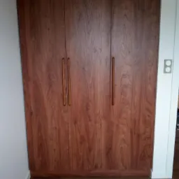 Trzydrzwiowa szafa wnękowa z okładziną imitującą drewno wiśniowe i długimi, cylindrycznymi uchwytami, dopasowana kolorystycznie do podłogi.