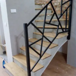 Betonowe schody z drewnianymi stopniami i czarną, geometryczną balustradą na tle ceglanej ściany.