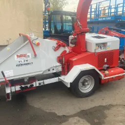 Profesjonalny rębak do gałęzi Bugnol BV. E10 w kolorze czerwono-białym, z podłączonym traktorem Kubota, na placu budowy, w tle podnośnik Genie GS-5390.