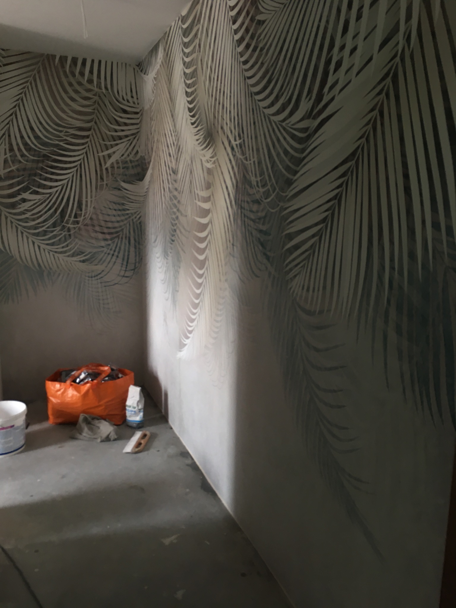 Ściana w pokoju z wzorem liści palmowych, widoczne narzędzia malarskie i wiadro z farbą, surowy beton na podłodze.