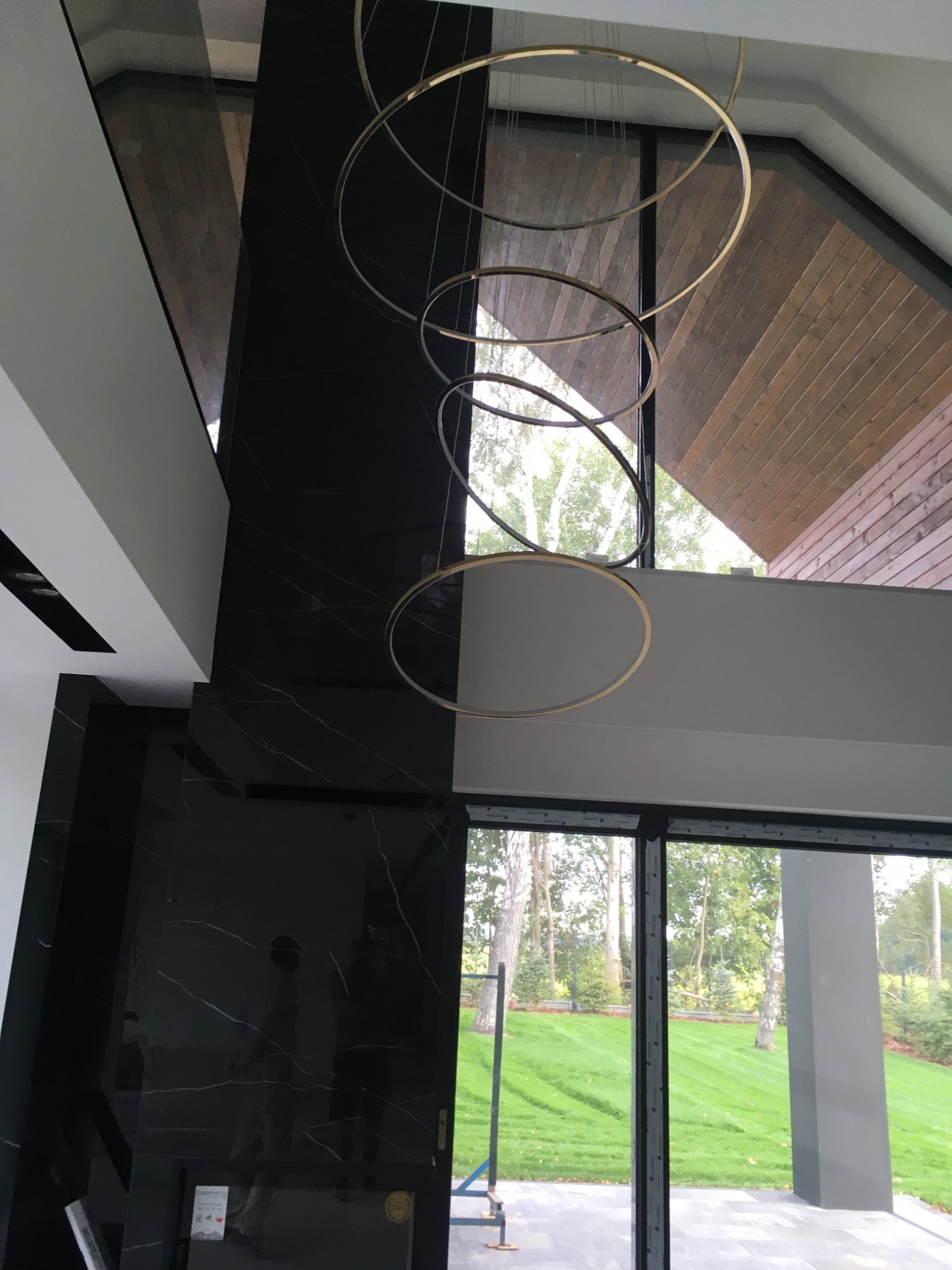 Wysokie wnętrze z czarną ścianą z marmuru, drewnianą ścianą pod skosem, dużymi oknami z widokiem na trawnik i nowoczesną lampą wiszącą z okręgów.