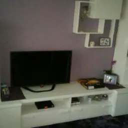 Biała, nowoczesna meblościanka z witrynami i szafkami RTV na tle fioletowej ściany, z elementami dekoracyjnymi na półkach.