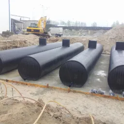 Cztery czarne, cylindryczne zbiorniki zakopane w ziemi na placu budowy w Mielcu, z widoczną koparką JCB w tle i przygotowanym wykopem.