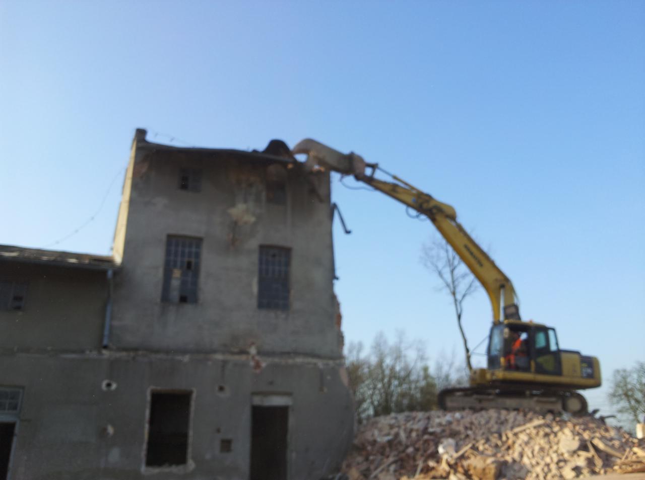 Żółta koparka z długim ramieniem rozbiera stary, szary, dwupiętrowy budynek z powybijanymi oknami, na tle błękitnego nieba.