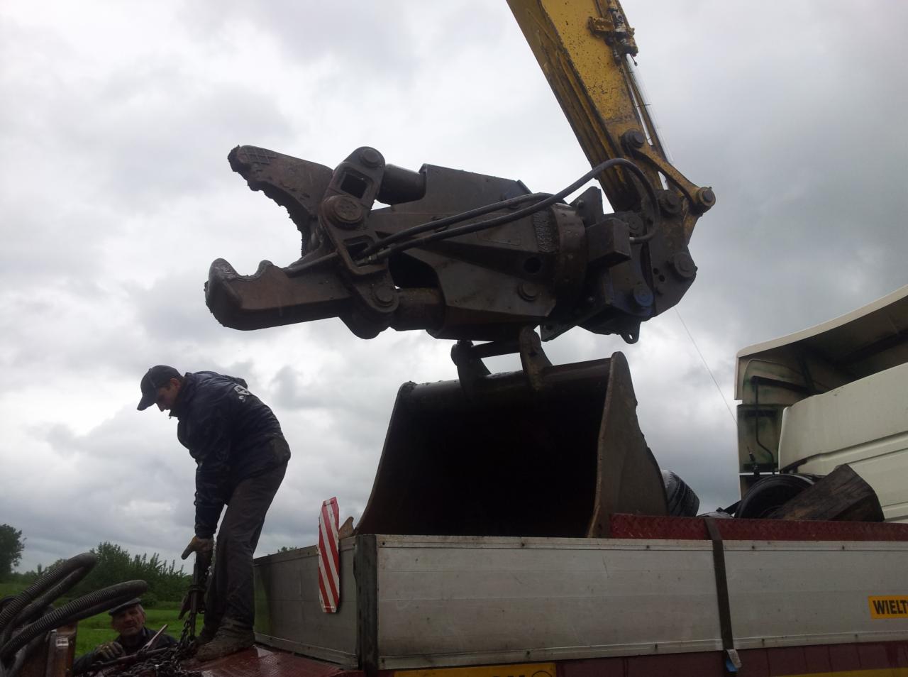 Operator w czarnej kurtce zabezpiecza łańcuchem łyżkę koparki z hydraulicznym chwytakiem na platformie ciężarówki, pochmurne niebo w tle.