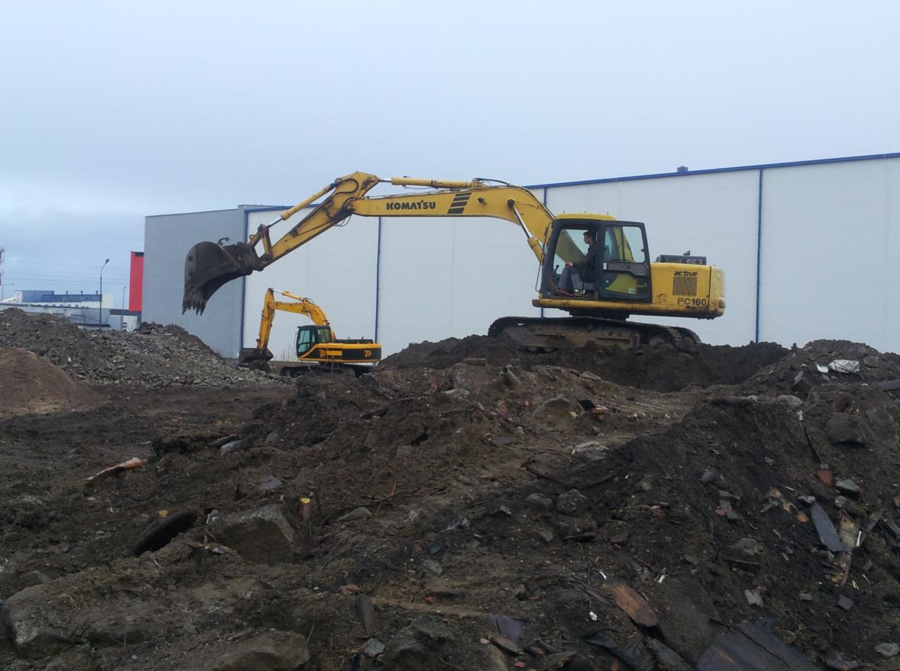 Dwa żółte koparki Komatsu na placu budowy, z widocznym operatorem w jednej maszynie, pracują na nierównym terenie z gruzem i ziemią na tle białej hali.