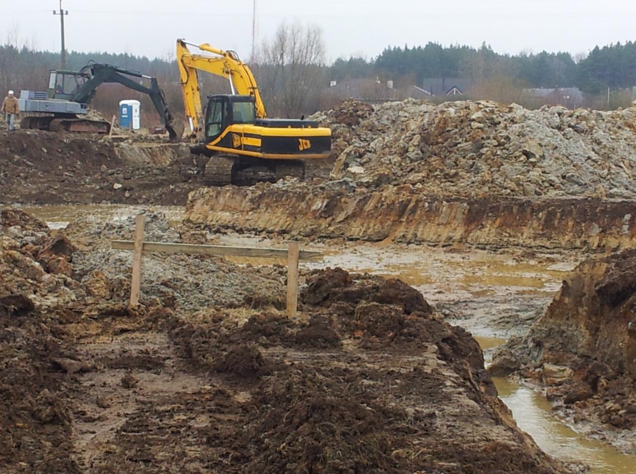 Żółta koparka JCB i szara koparka pracują na placu budowy z błotnistym terenem, widoczny przenośny wychodek i sterta ziemi w tle.