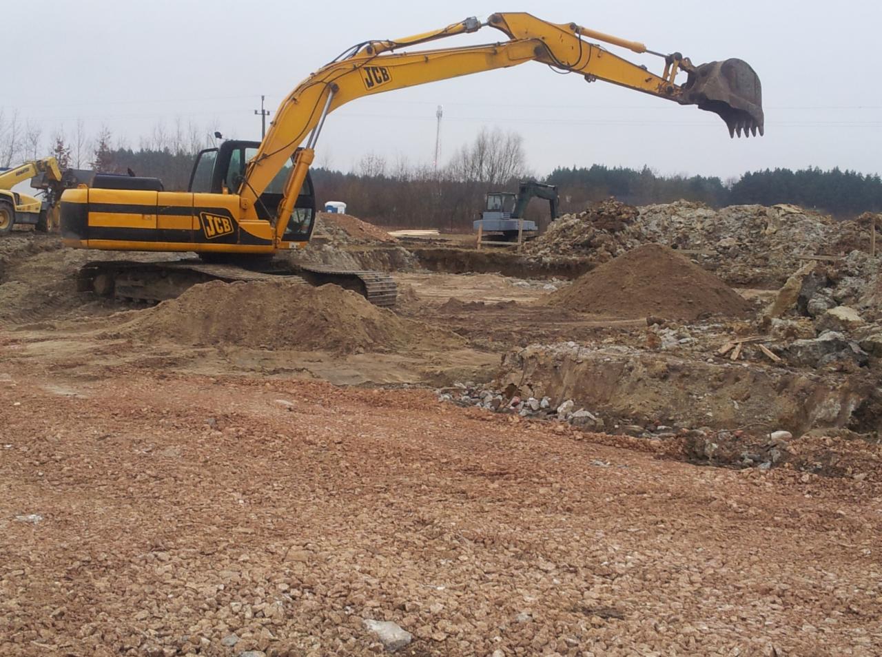 Żółta koparka JCB na placu budowy, wykopane rowy i hałdy ziemi, w tle widoczny las i słup wysokiego napięcia.