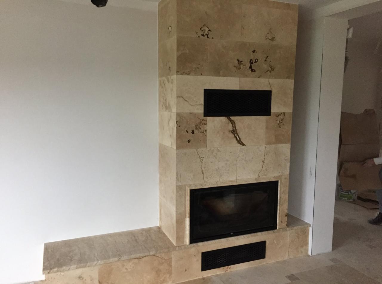 Obudowa kominka z trawertynu z czarnym wkładem, widoczne kratki wentylacyjne, surowa ściana i podłoga w trakcie wykańczania.