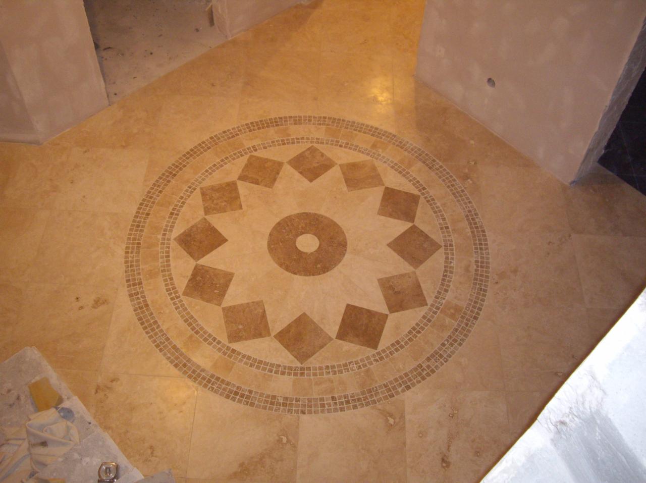 Beżowa podłoga z trawertynu z dekoracyjnym, okrągłym wzorem z mozaiki w kształcie gwiazdy i obramowaniem z drobnych kostek, widoczne fragmenty ścian w stanie surowym.