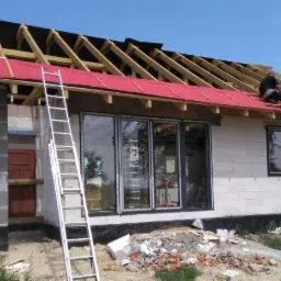 Budowa domu jednorodzinnego w trakcie: widoczne ściany z bloczków, zamontowane okna, oraz konstrukcję dachu z deskami i czerwoną membraną, dwóch pracowników na dachu.