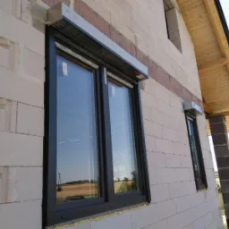 Nowe okna z szarą ramą i zamontowanymi roletami zewnętrznymi na ścianie budynku z betonu komórkowego w trakcie budowy.