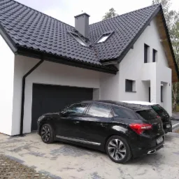 Nowoczesny dom jednorodzinny z czarną bramą garażową, antracytową dachówką i dwoma samochodami na podjeździe wyłożonym ażurowymi płytami.