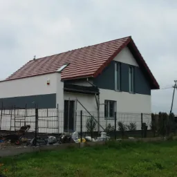 Dwukondygnacyjny dom jednorodzinny z nową, czerwoną dachówką i świeżo pomalowaną elewacją w kolorze białym i szarym, otoczony metalowym ogrodzeniem i zielonym trawnikiem, widok z zewnątrz...