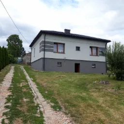 Dwukondygnacyjny dom z szarą elewacją i dekoracyjnymi pasami, widoczny z perspektywy podjazdu z ceglaną ścieżką i zielonymi tujami po lewej stronie.