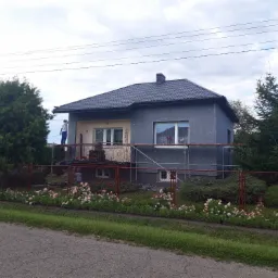 Dom jednorodzinny w trakcie prac elewacyjnych: szara elewacja z widocznymi płytami izolacyjnymi, fragment ocieplonej ściany w kolorze beżowym, pracownik na rusztowaniu przy balkonie, nowy dach...