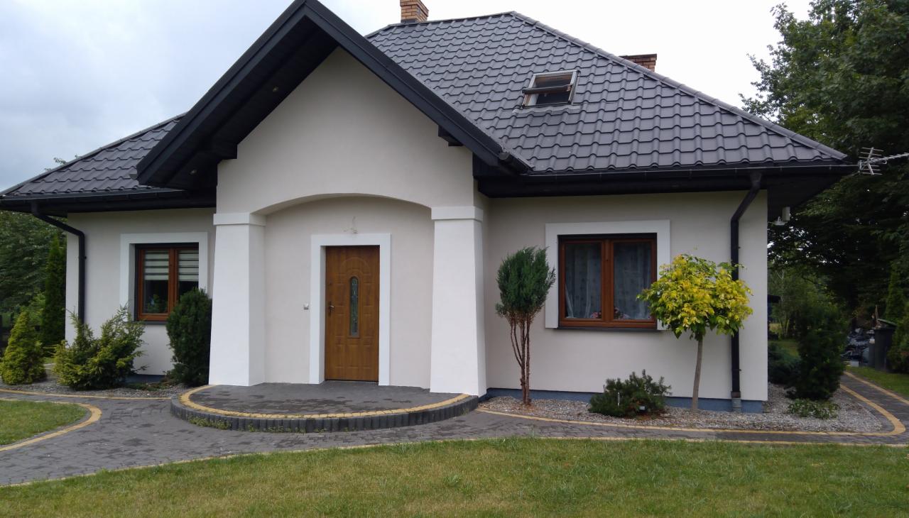 Dom jednorodzinny w Zamościu po tynkowaniu elewacji, z ciemną dachówką i jasną elewacją, widok z przodu na wejście i zadbany trawnik.
