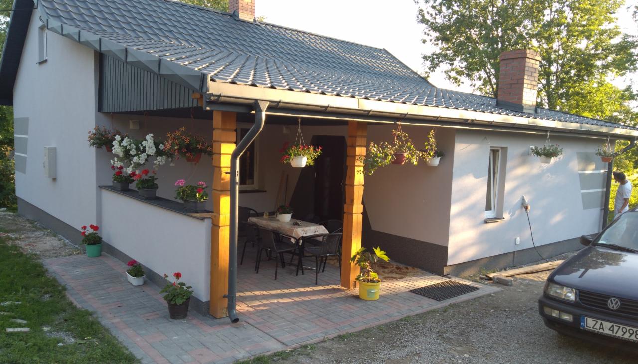 Wykończony dom z nową szarą elewacją, ciemną dachówką i zadaszonym tarasem z drewnianymi filarami, ozdobiony wiszącymi i stojącymi doniczkami z kwiatami.