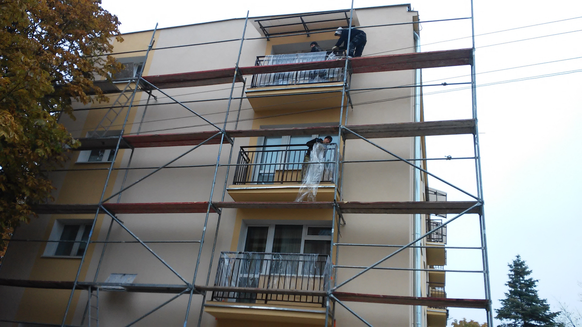 Ocieplanie elewacji budynku wielorodzinnego z balkonami, widoczne rusztowania i pracownicy zabezpieczający balkony folią ochronną.