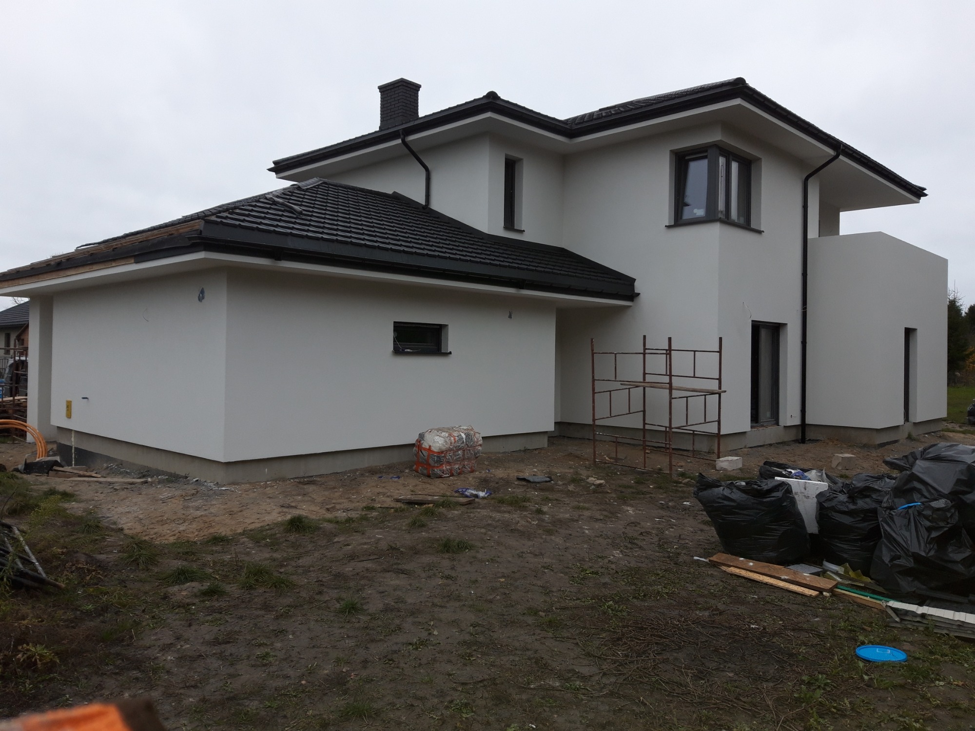 Dwukondygnacyjny dom z białą elewacją i czarnym dachem, widoczne rusztowanie, teren wokół domu w trakcie prac wykończeniowych, worki z odpadami budowlanymi, częściowo wykończony dach.