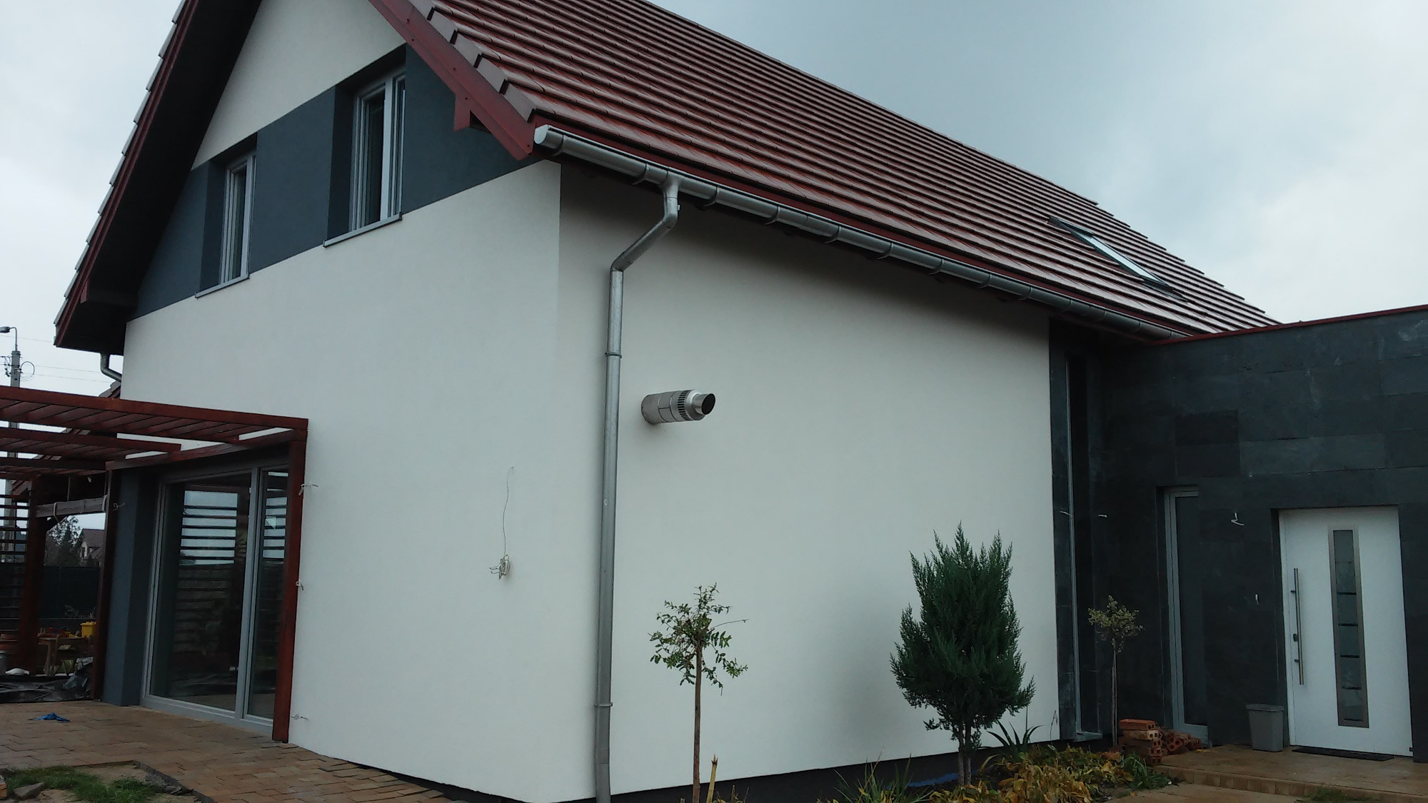 Wykończona elewacja domu jednorodzinnego z jasnym tynkiem, ciemnoszarą wstawką pod dachem, czerwoną dachówką i grafitową ścianą przy wejściu, widoczne rynny i wylot wentylacji.