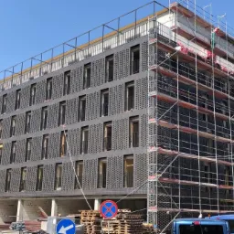 Wielopiętrowy budynek z ciemną elewacją w trakcie prac wykończeniowych, widoczne rusztowania i palety z materiałami budowlanymi, znak drogowy w tle.