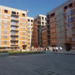 Wielopiętrowy budynek mieszkalny w trakcie budowy, z widocznymi cegłami, betonowymi elementami konstrukcyjnymi i banerami reklamowymi na elewacji, sfotografowany z poziomu placu wyłożonego kostką...