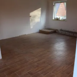 Ułożona podłoga z brązowych płytek imitujących drewno w pustym pokoju, widoczne niedokończone ściany, okno z parapetem i drabina aluminiowa.