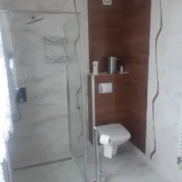 Nowoczesna łazienka z kabiną prysznicową typu walk-in, toaletą i elementami wykończenia w drewnie, akcentująca marmurowe płytki podłogowe i ścienne z delikatnym wzorem.