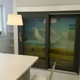 Nowoczesne, przesuwne drzwi tarasowe z ciemną ramą i fototapetą przedstawiającą krajobraz polny z chmurami, widoczne z wnętrza jasnego pomieszczenia z białymi ścianami, stołem i oknem.