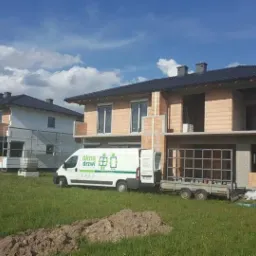 Dwa domy w stanie surowym otwartym z zamontowanymi oknami, na trawniku stoi biały bus z logo firmy montującej okna i drzwi, do busa doczepiona przyczepka z ramą okienną.