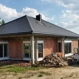 Surowy stan domu jednorodzinnego w Lubinie z zamontowanymi oknami i roletami zewnętrznymi, widoczny stos cegieł i samochód dostawczy firmy.