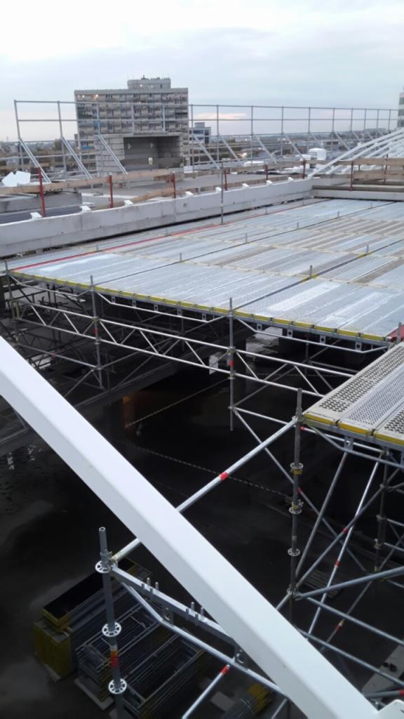 Widok z góry na rozległe rusztowanie na dachu budynku w trakcie budowy, z widocznym w tle budynkiem mieszkalnym i pochmurnym niebem.