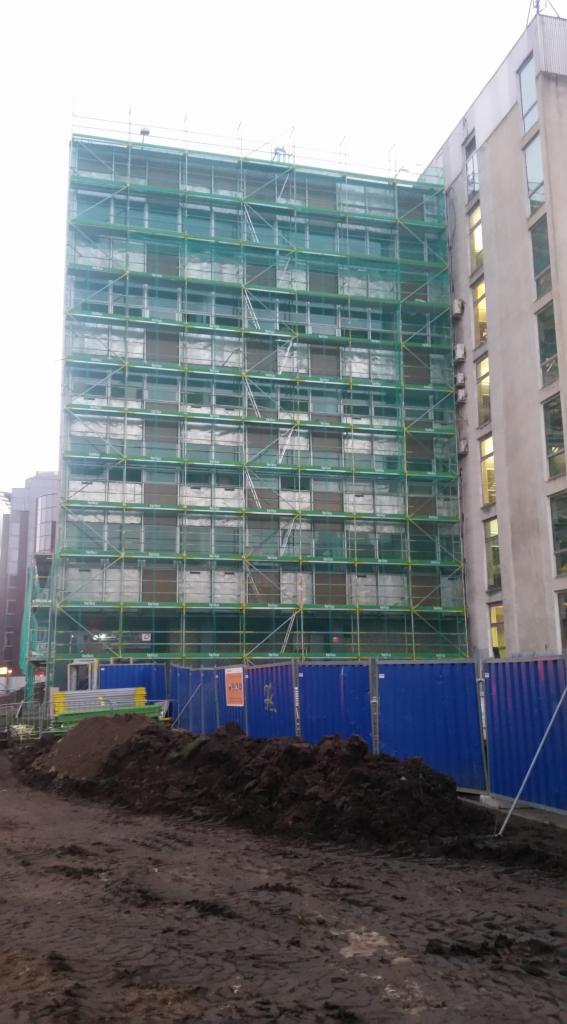 Wielopiętrowy budynek w trakcie remontu, otoczony rusztowaniami z zieloną siatką ochronną i zabezpieczony niebieskim ogrodzeniem tymczasowym na placu budowy z widoczną stertą ziemi.