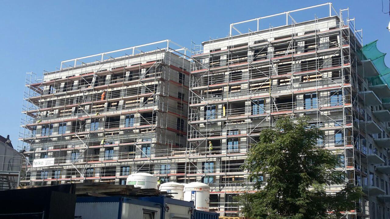Wielopiętrowy budynek w trakcie budowy, otoczony rusztowaniami, z widocznymi pracownikami w odblaskowych ubraniach i zbiornikami na dole.